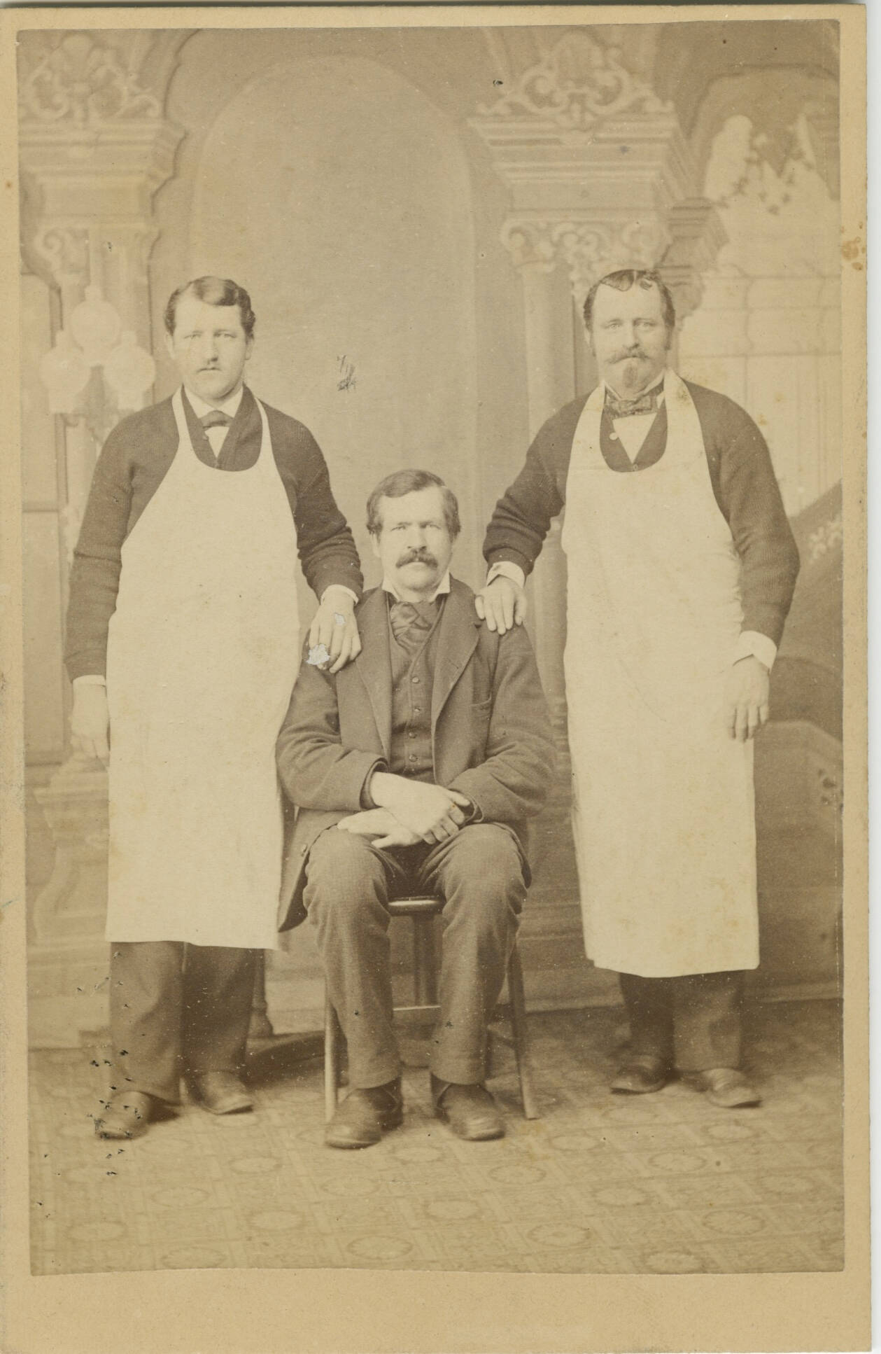 Black and white cabinet card photograph of three men, the middle one is seated and the other two are wearing aprons.